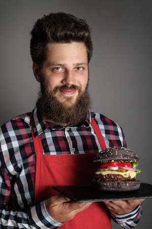 Male cook in red apron giving you fresh self made black burger and smilingの写真素材