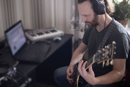 Bearded man musician playing music and composing a song with electric guitar piano and laptop...