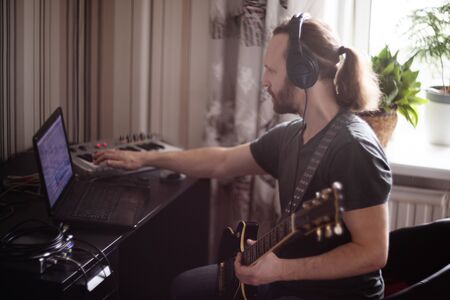 Bearded man musician playing music and composing a song with electric guitar piano and laptop...