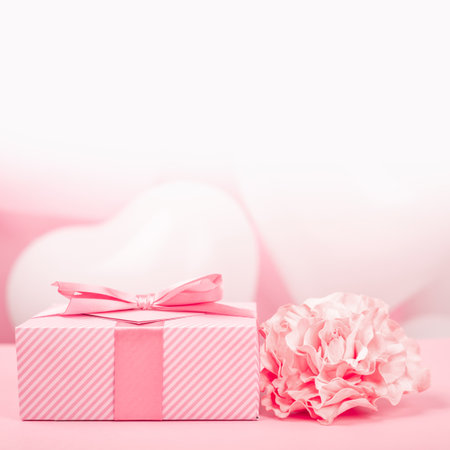 Valentine Day gift in a box wrapped in striped paper and tied with silk ribbon bow and heart shaped greeting card on pink balloons background with copy space for textの写真素材