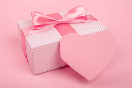 Valentine Day gift in a box wrapped in striped paper and tied with silk ribbon bow and heart shapes greeting card on pink background with copy space for textの写真素材