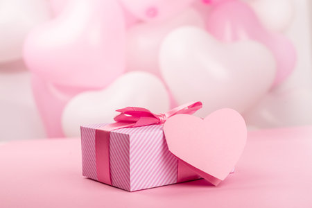 Valentine Day gift in a box wrapped in striped paper and tied with silk ribbon bow and heart shaped greeting card on pink balloons background with copy space for textの写真素材