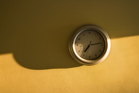 Analogue clock on the yellow wall showing 7:14 time, light from the side, long shadows emphasising early morning sun.の写真素材