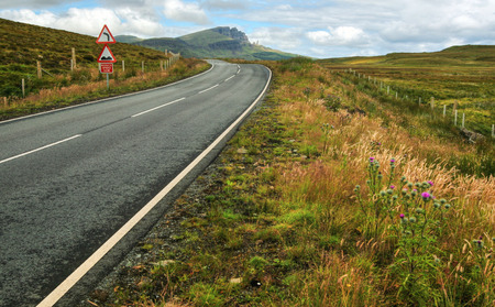 Wide shot of empty road, curve, bumps and reduce speed now sign with Old Man of Storr mountain in...