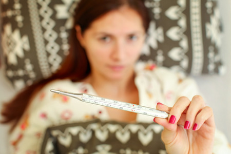 Sick young woman lying in the bed, covered with duvet,  holding mercury thermometer showing...