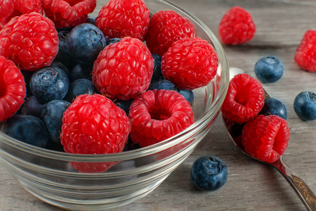 Small glass bowl of fresh raspberries and blueberries with spoon next to it, placed on gray wooden...