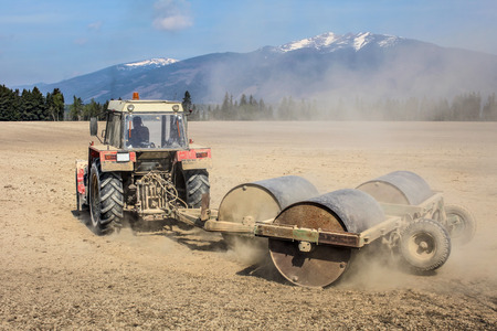 Tractor pulling heavy metal rollers on a dry field with mountains in background. Spring ground...