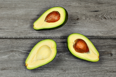 Tabletop view - three halves of cut avocado, some with seed pit, on gray wood desk.