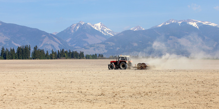 Tractor pulling rollers over dry fields in spring, mountains with little snow in background.