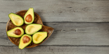 Tabletop view - avocado cut in half, arranged on wooden bowl, with dark grey wood desk under. Banner...