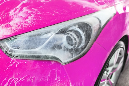 Front headlight of pink car washed in self serve carwash, water spraying leaves white foam on glassの写真素材