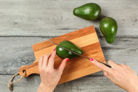 Tabletop view, woman hands holding knife, cutting green whole avocado on chopping board.の写真素材