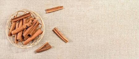 Small wicker basket with cinnamon bark sticks on linen tablecloth, view from above. Wide banner...