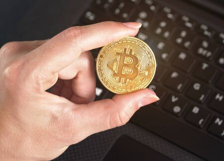 Woman hand holding one golden coloured bitcoin coin over black laptop keyboard - cryptocurrency...