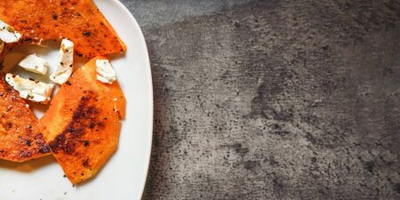 Grilled orange butternut squash pieces with feta cheese on white plate, dark stone like board under...