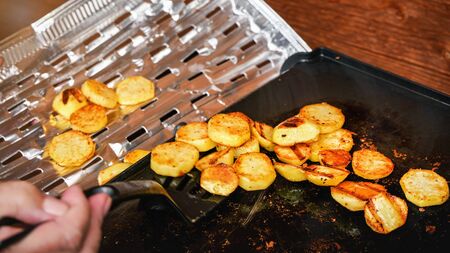 Potatoes cut to small circles, seasoned with spices, grilled on electric grill - woman hand holding spatula moving cooked food onto aluminium foil tray.の写真素材
