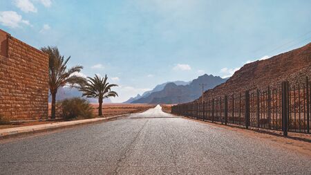 Long straight road with desert hills in distance, brick wall and iron fence on sides. Scenery at the...