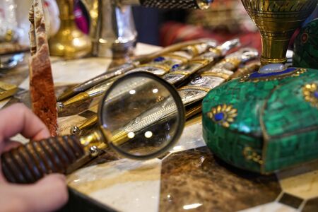 Examining details on traditional daggers through loupe magnifying glass in vintage antiques shop,...
