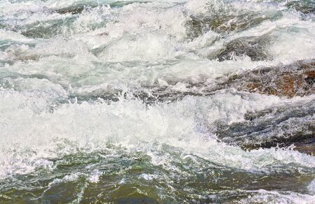 Rapid spring river flowing over rocks on sunny day, forming white water waves, closeup detail -...
