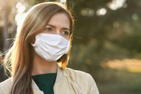 Young woman wearing white cotton virus mouth nose mask, blurred sunset lit trees in background,...