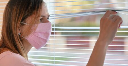 Young woman in pink home made cotton virus face mask, looking through window blinds outside....