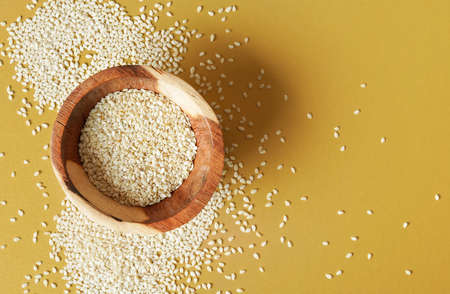Tiny white sesame seeds in small wooden bowl, on yellow desk, view from aboveの写真素材