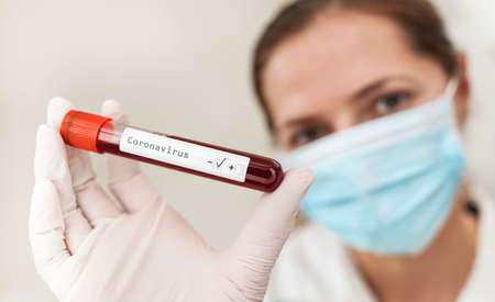 Hand in white gloves holding vial with blood, label coronavirus test, negative result, blurred woman in blue mouth mask face background. Covid 19 testing conceptの写真素材