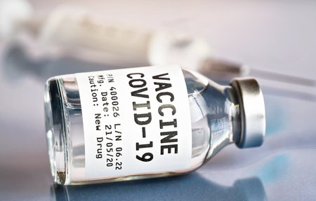 Glass bottle with Covid-19 vaccine (own sticker, dummy data, not real product), blurred hypodermic syringe needle near, closeup detail - coronavirus cure conceptの写真素材
