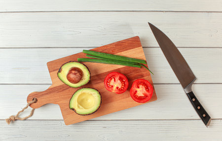 Avocado halves, tomato, spring onions - basic guacamole ingredients on working board, knife near, view from aboveの写真素材