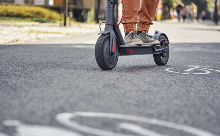 Young man in casual trousers riding an electric scooter detail on his feet and wheel over asphalt...