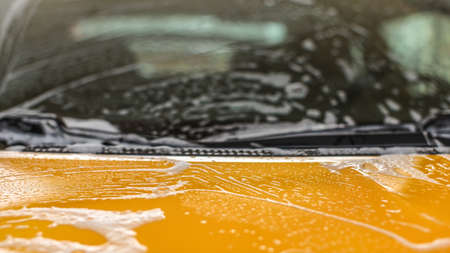 Closeup detail - front hood of yellow car covered with white shampoo foam when washed in carwash. Abstract automotive cleaning background with space for text.の写真素材