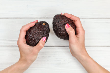 Top down view, woman hands with pink nails holding two whole dark ripe avocados over white boards...