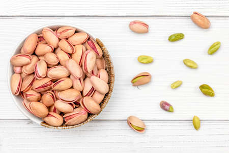 Overhead shot, roasted pistachios nuts in small bowl, some scattered on white boards desk.の写真素材