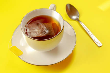 Porcelain cup with hot tea, bag just brewing in, silver spoon next on yellow board, lit by morning...