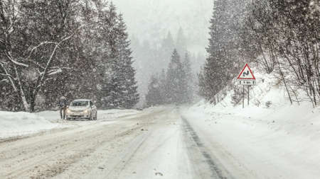 Driving through heavy snowstorm blizzard at forest road, warning curves ahead sign right, and...