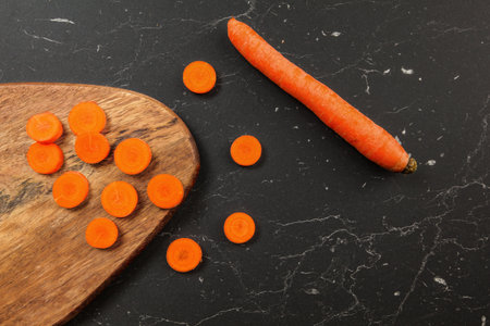 Carrot sliced to small circles and chopping board, whole one next to it on black working desk, photo...