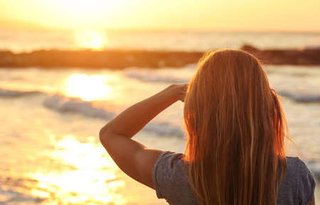 Sporty woman watches sunset over the sea, shading her eyes with one hand, view from behind only hair visibleの写真素材