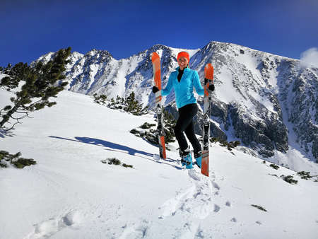 Young woman in light blue jacket standing, smiling happy holding with mountaineering ski and poles in hands, sun shines on snow, bright sky over mountains behindの写真素材