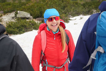 Young woman hiker in red jacket and sunglasses smiling to two other hikers, snow and trees behind...
