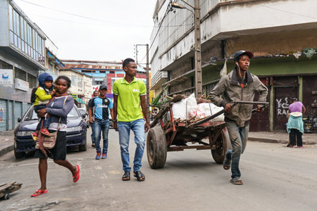Antananarivo, Madagascar - April 24, 2019: Unknown Malagasy man pulling cart with white bags, woman with child on her hands walking near, more locals around.のeditorial素材