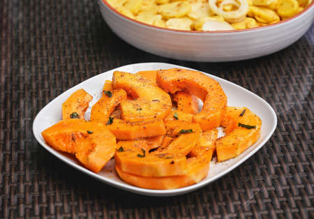 Grilled orange butternut squash pieces with herbs, on white square plate, another full of potatoes in backgroundの写真素材