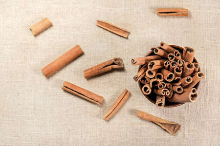 Closeup detail - Cinnamon bark sticks in small wooden cup, some scattered on linen tablecloth view...