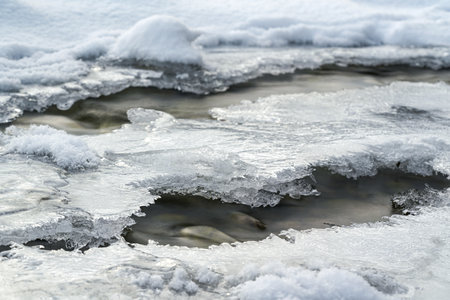 Shallow forest river covered with crystal snow and ice layer in winter, white patches over stones, long exposure photoの写真素材