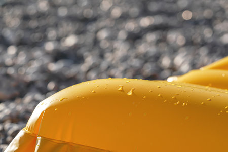 Sun shines to yellow coloured inflatable with drops on water on surface, blurred rocky beach background, closeup detailの写真素材