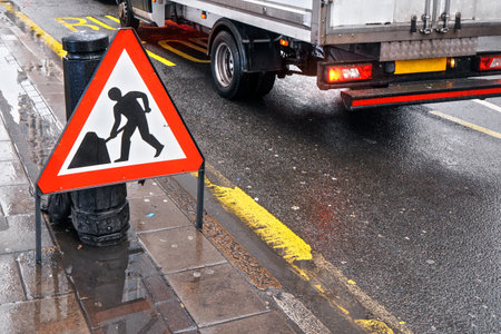 Warning roadworks sign at wet road next to pavement, van moving in background over Bus Stop writing...