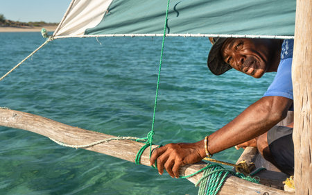 Anakao, Madagascar - May 03, 2019: Unknown Malagasy fisherman looking back under main green sail of his piroga (small fishing boat), green blue sea during sunny day in backgroundのeditorial素材