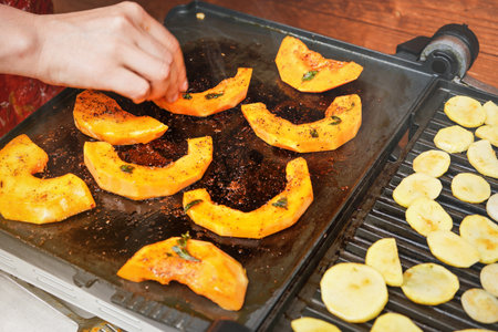 Bright orange butternut squash pieces grilled on electric grill, blurred woman hand moving pieces of vegetables over hot plateの写真素材