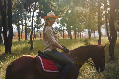 Young woman in shirt and straw hat, riding brown horse in the park, blurred grass and trees backgroundの写真素材