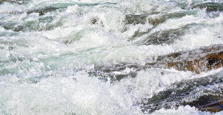 Rapid spring river flowing over rocks on sunny day, forming white water waves, closeup detail -...