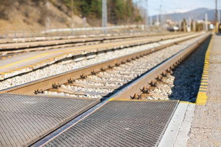 Empty train station in small city on sunny day, shallow depth of field photo, focus on concrete platform, passenger crossing, and steel rail foregroundの写真素材